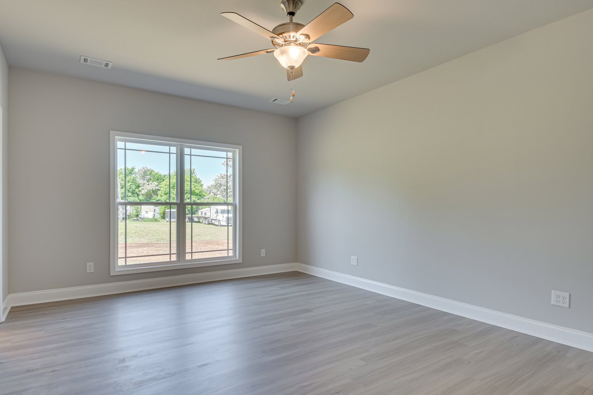 Room with light wood flooring, white walls, ceiling fan with light fixture, window overlooking field and trees, white electrical outlet on wall, simple molding along baseboards.