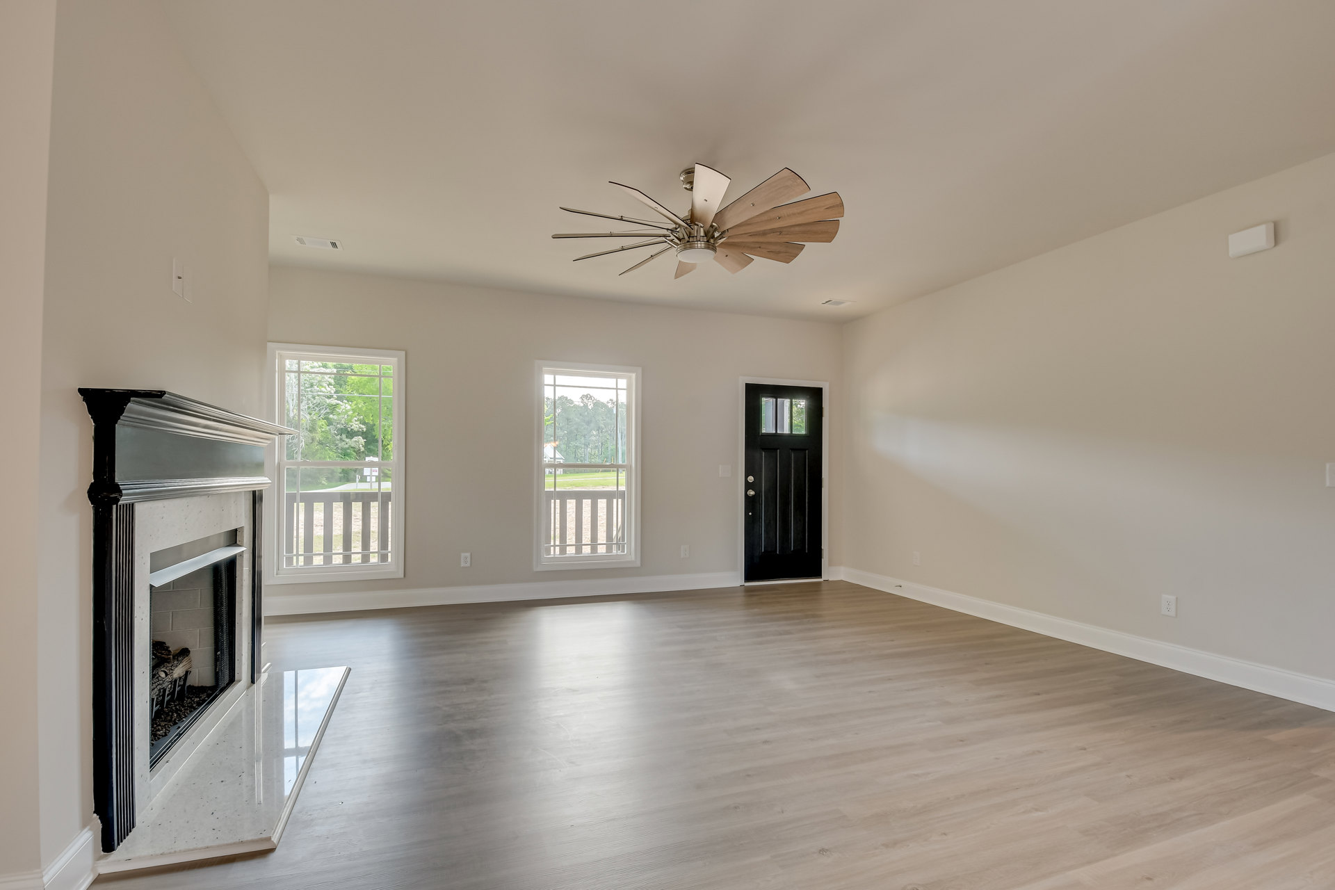 Living room with stone fireplace, black door with glass window, wood flooring, ceiling fan with light, large window showing trees outside
