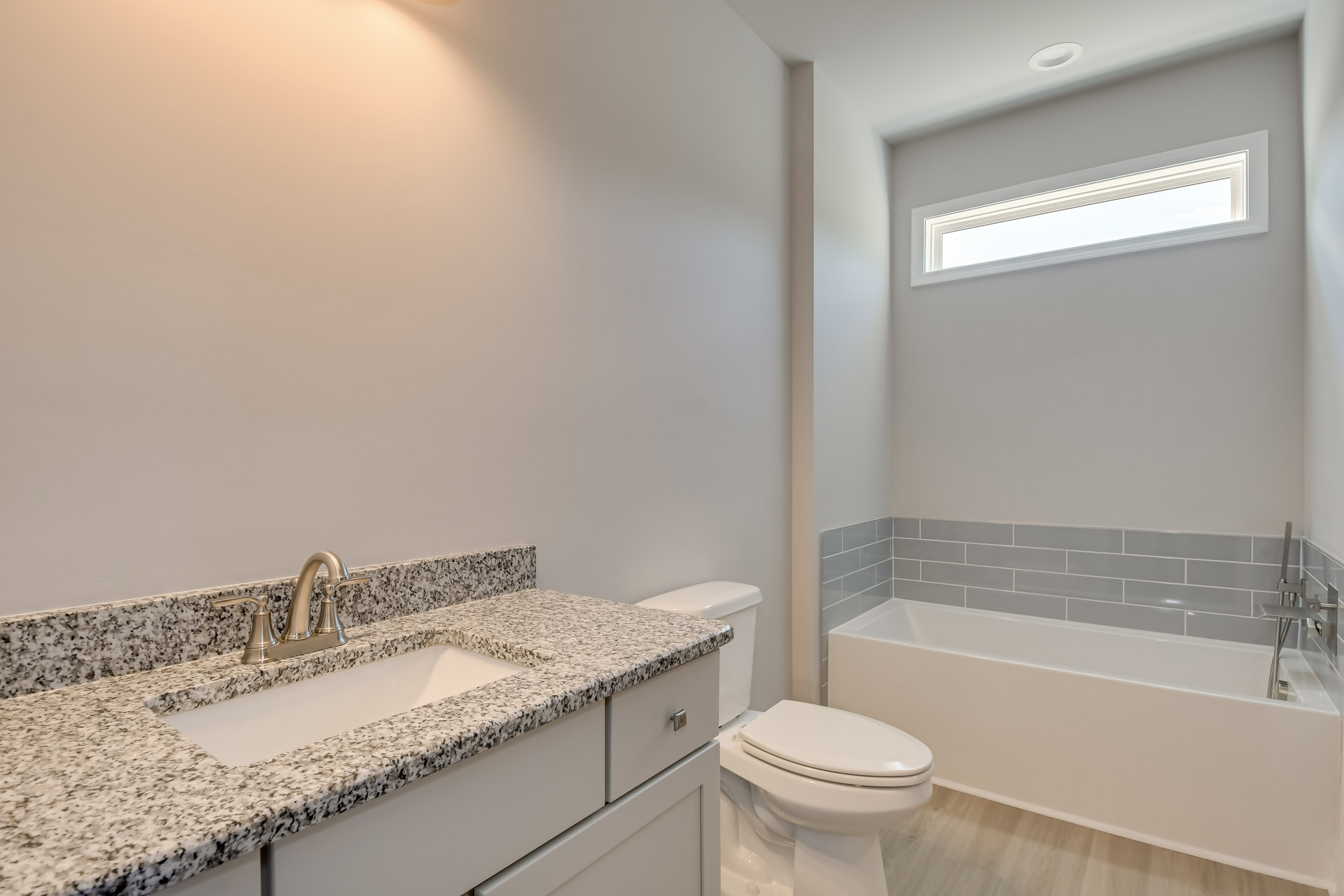 Marble countertop vanity with chrome faucet, white undermount sink, freestanding white bathtub beside wood flooring, wall-mounted mirror, white toilet, and recessed ceiling light.