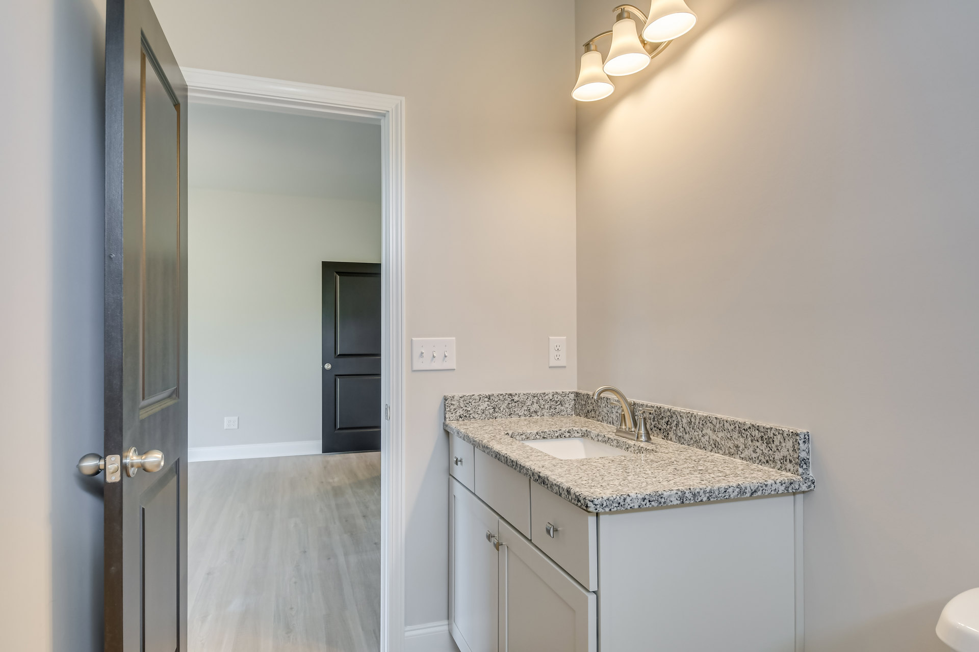 Marble countertop with undermount sink, white cabinetry, three-light fixture above mirror, wood flooring, black and white accent wall, close-up of light switch and door