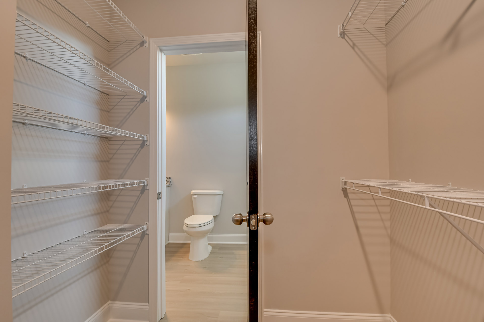 White toilet with closed lid beside white wire shelves mounted on plaster wall, tile floor, and chrome door knob visible in foreground.