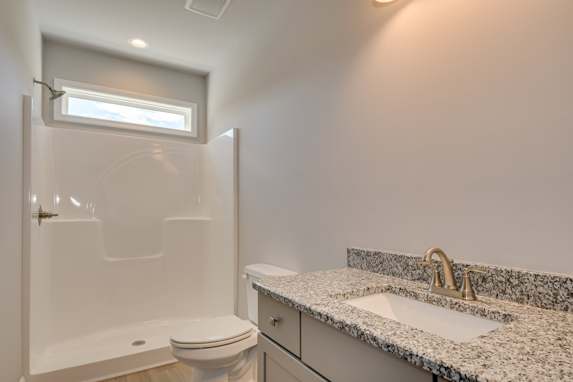 Modern bathroom with white bathtub beneath a window, single sink vanity with chrome faucet, wall-mounted mirror, and white toilet on light tile flooring