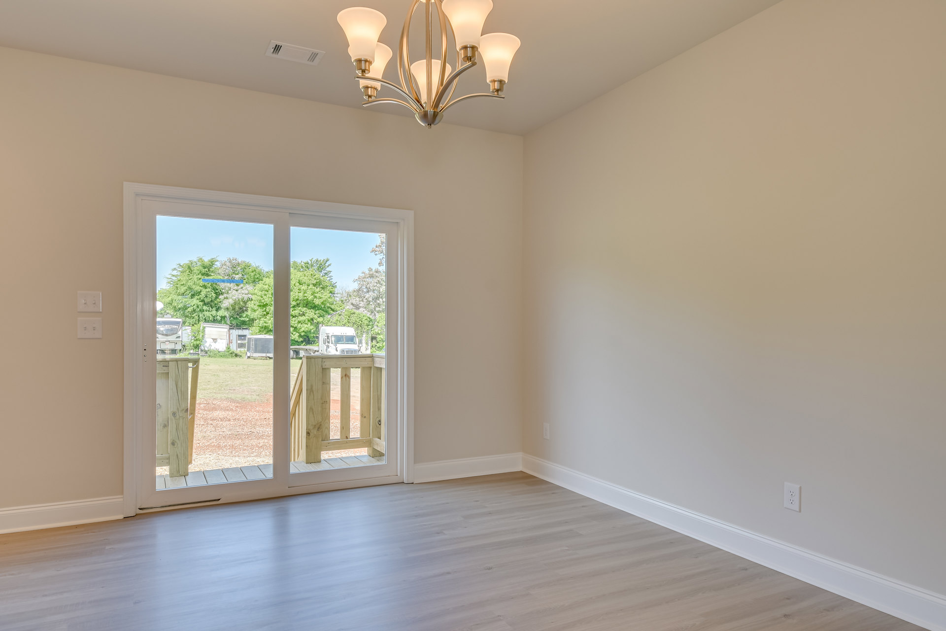 White wood flooring in a bright room featuring a white door, sliding glass door opening to a deck with wooden railing, and a modern chandelier hanging from a plaster ceiling.
