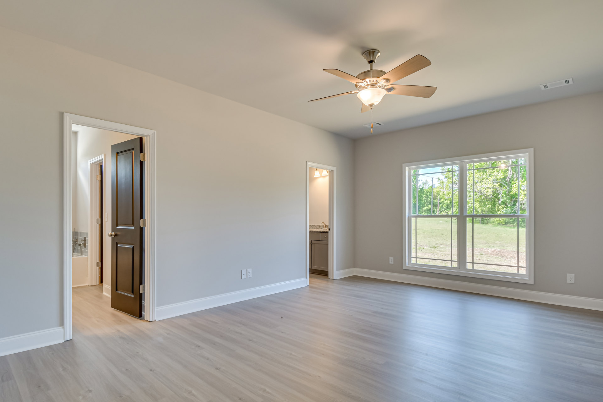 Bedroom with wood flooring, white walls, ceiling fan with light fixture, large window overlooking trees, door opening to bathroom
