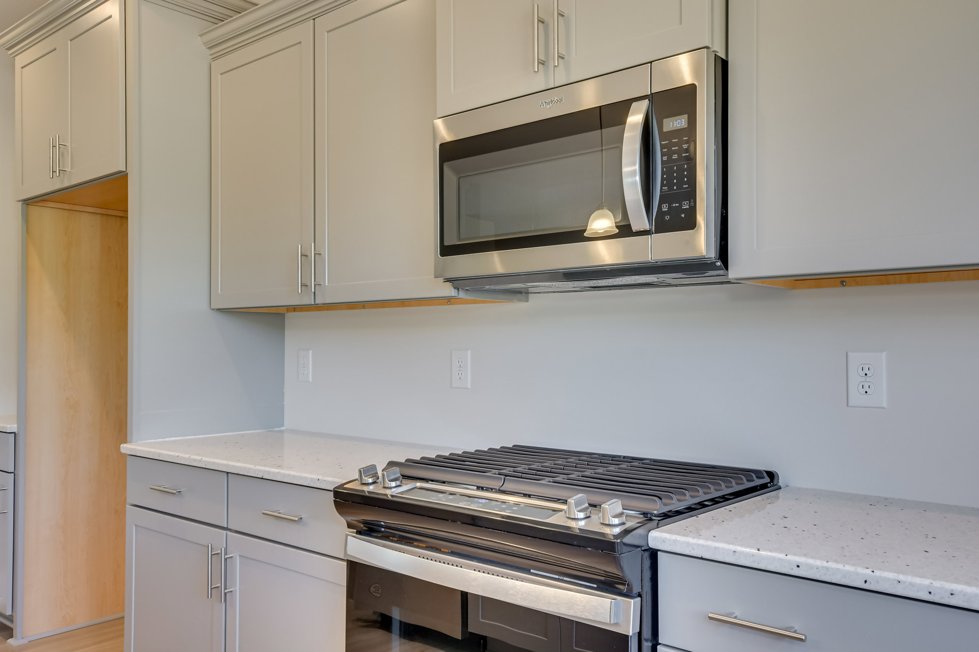 White shaker cabinets and quartz countertops surround a stainless steel microwave oven, with a gas stove below, subway tile backsplash, and a bell-shaped pendant light overhead.