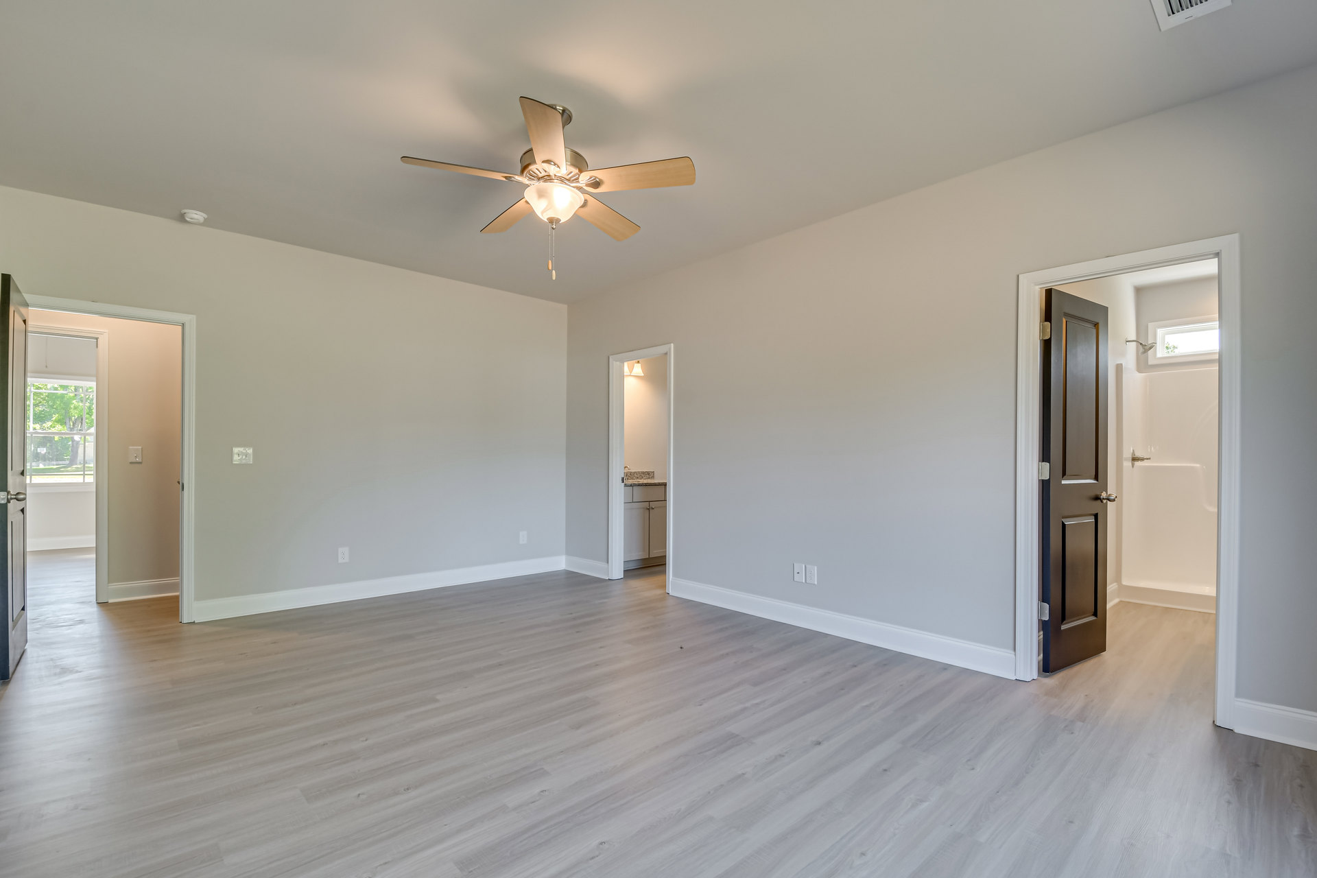 Wood flooring with white baseboards, ceiling fan with light fixture, white door open to bathroom, neutral walls and ceiling