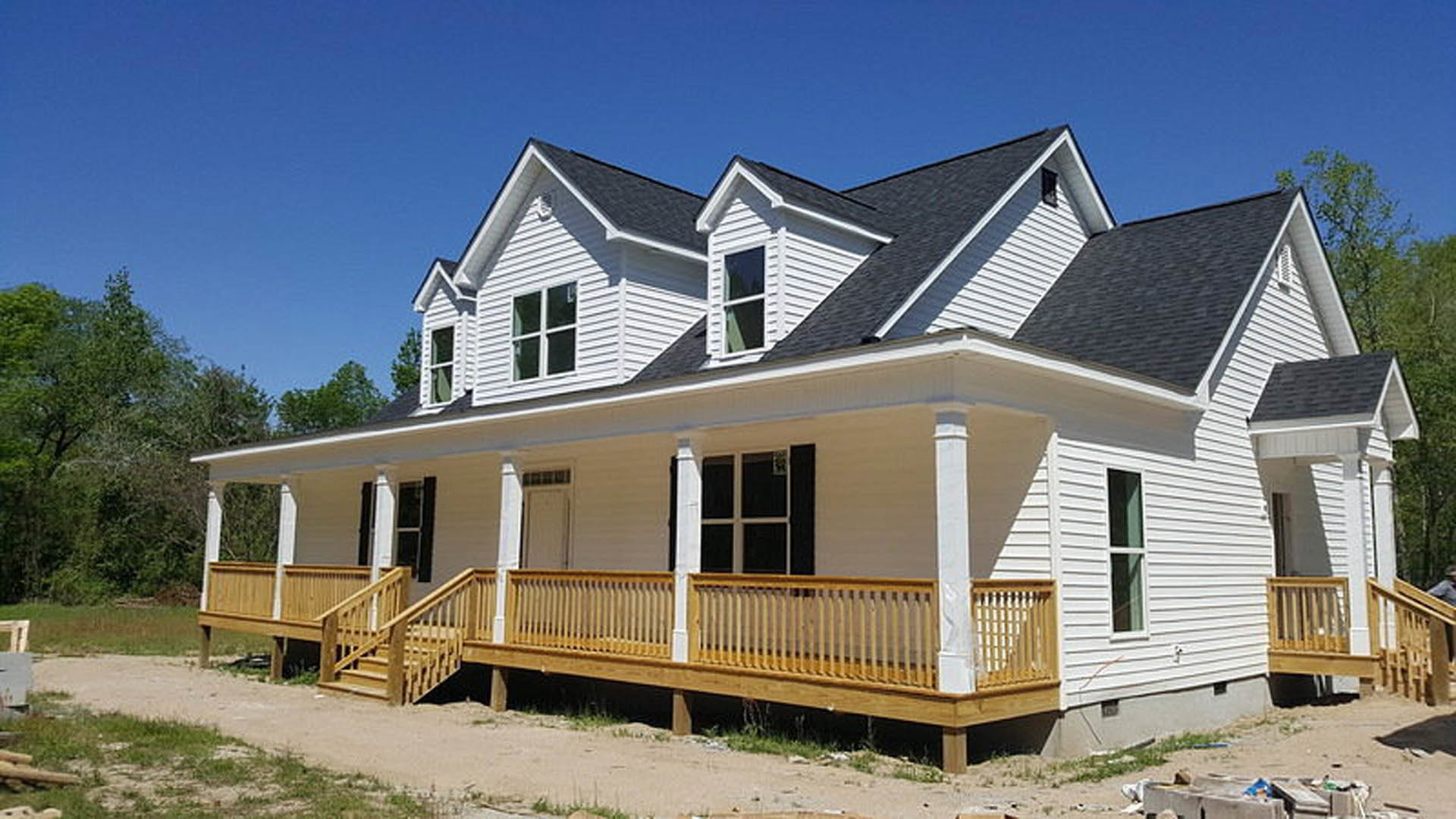 Front porch with wooden railing and white pillars, green and white framed windows, light siding, leafy tree nearby