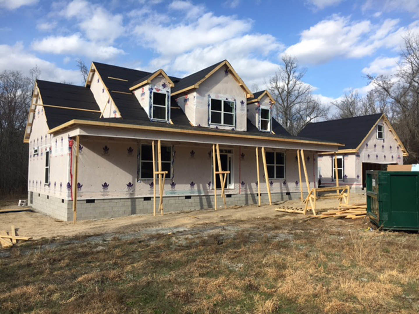 Wood-framed house under construction with exposed scaffolding, green metal container nearby, grassy yard with dirt patches, brick wall, and window detail beneath a cloudy sky