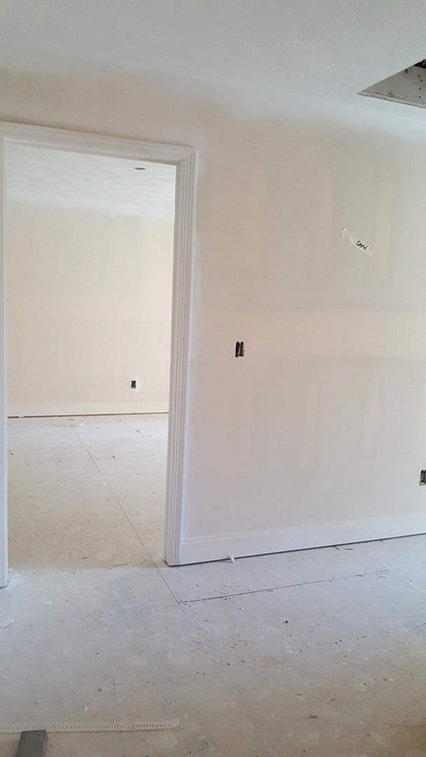 White-walled room with white ceiling, white door, and white floor; close-up of person's feet on smooth flooring; wall features a square opening.