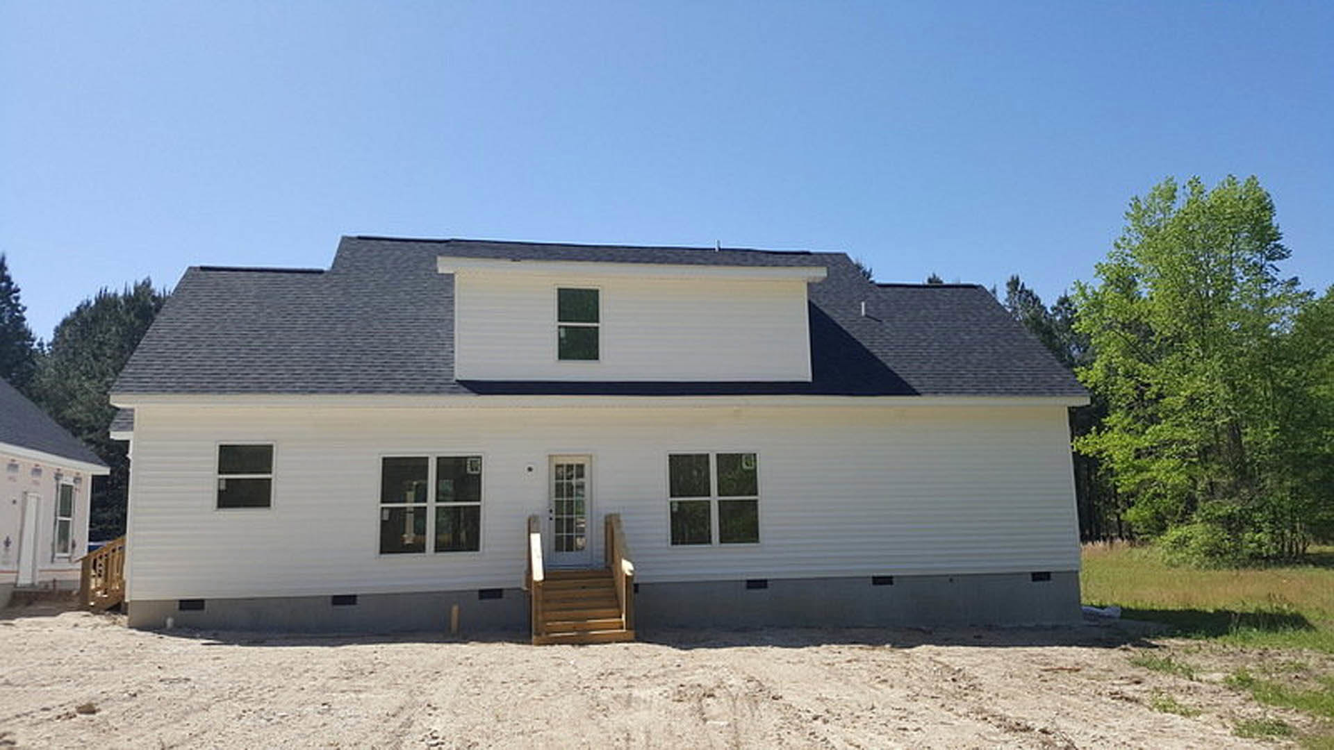 Two-story house with wooden staircase leading to a white door with glass panes, dirt yard in front, trees and sky in background, close-up window visible on facade