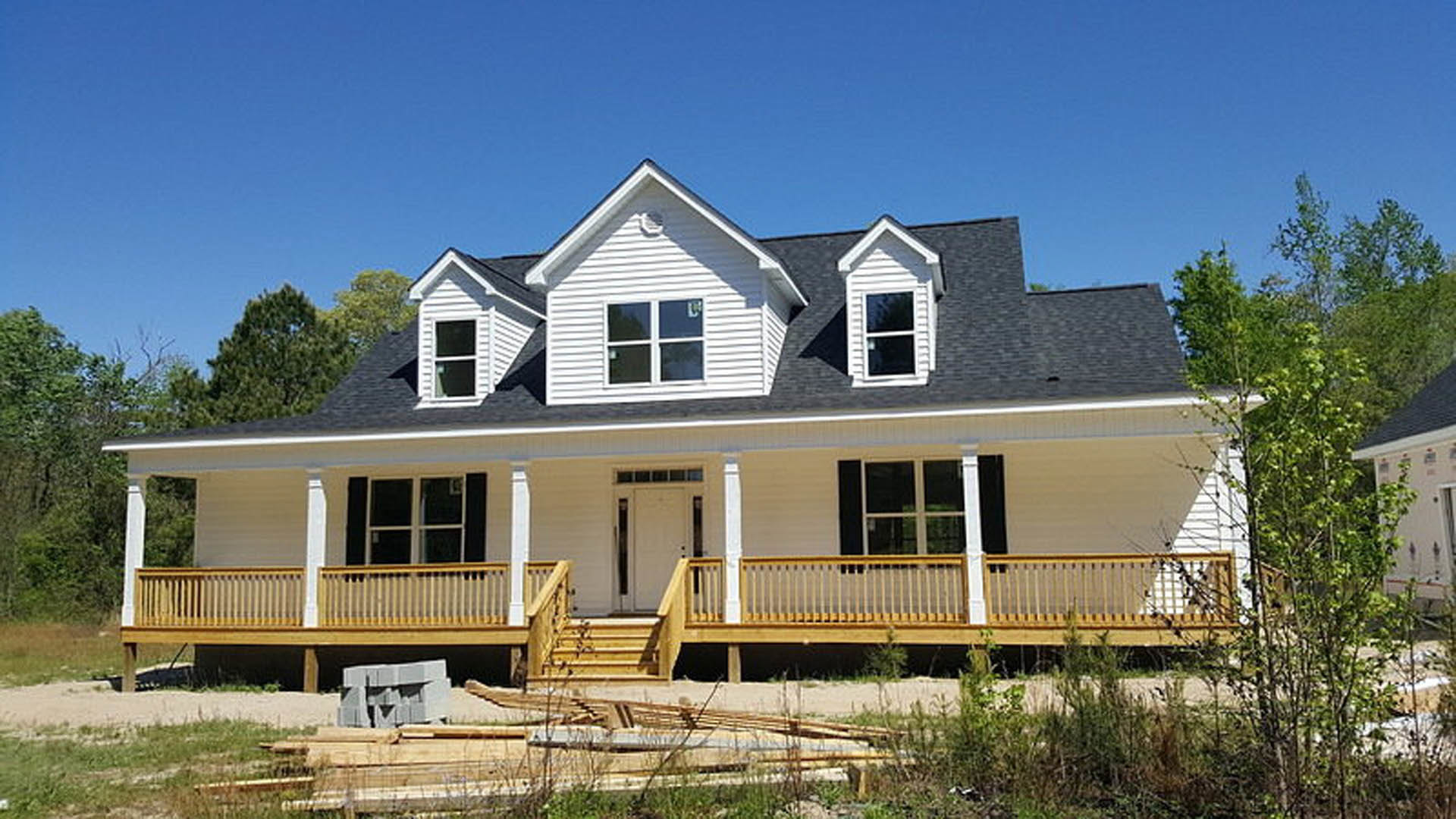 Two-story house with gray siding, wooden porch and deck, white door with glass panels, large windows, and surrounding trees