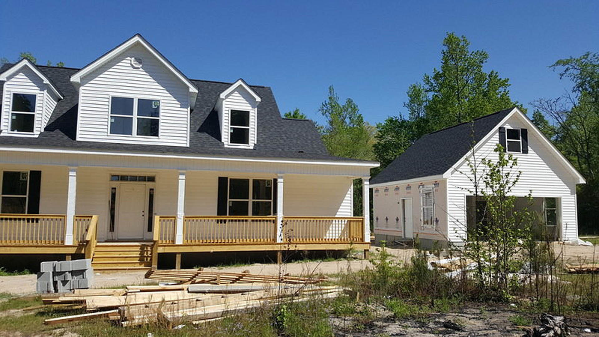 White house under construction with black shutters, white framed windows, porch, wooden deck with railing, surrounded by trees in the background