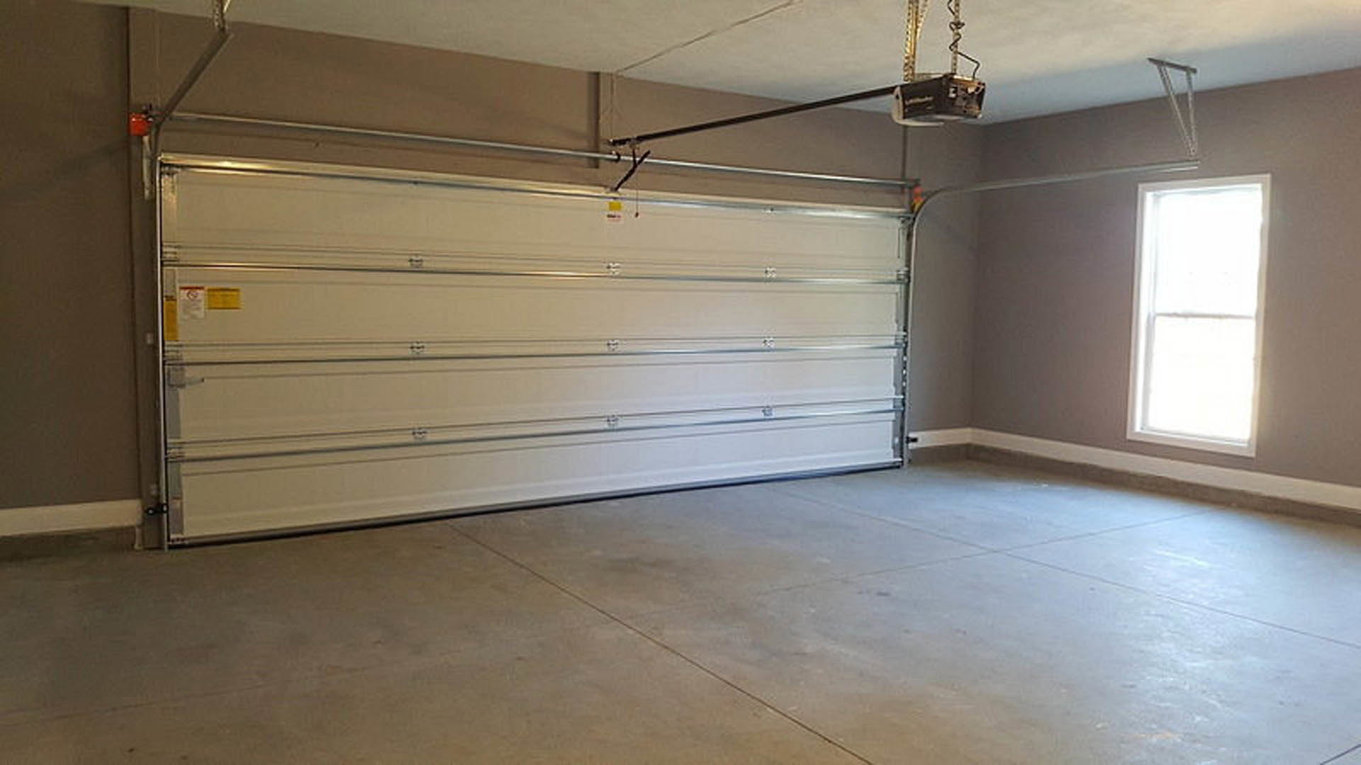 Garage interior with white metal door, concrete floor, plaster walls, ceiling, and a window; yellow rectangular object and bucket placed near the door.