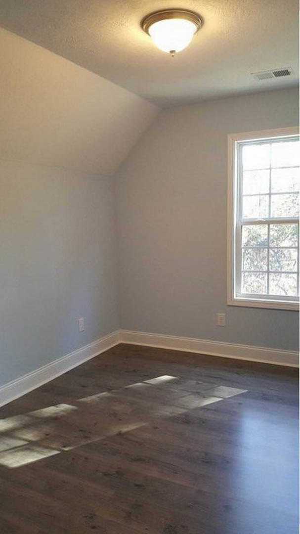 Sunlit wood floor beneath large window, white plaster walls, ceiling light fixture, simple molding along baseboards
