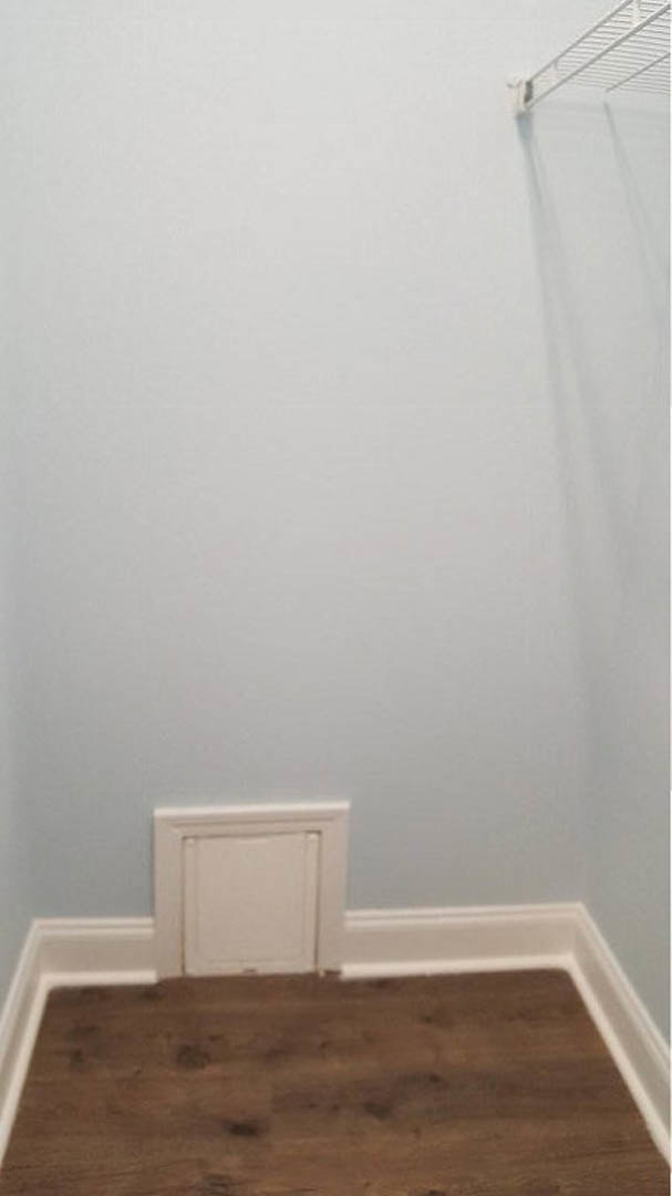 Small square white vent on a white wall above wooden floor with white trim, adjacent to a white door and metal ladder in an empty room.