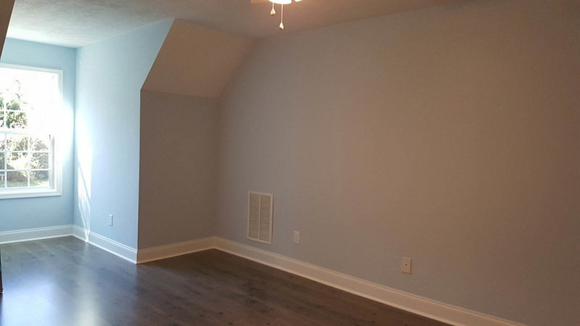 Wood flooring and white wall trim in a room with a ceiling fan, vent, white-framed window, and white wall outlet