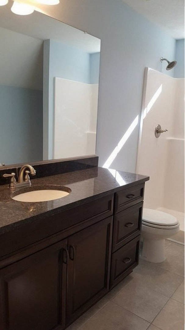 Modern bathroom featuring white ceramic sink with chrome faucet, matching toilet with closed lid, light gray tile walls, wood cabinetry with drawers, and stone countertop.