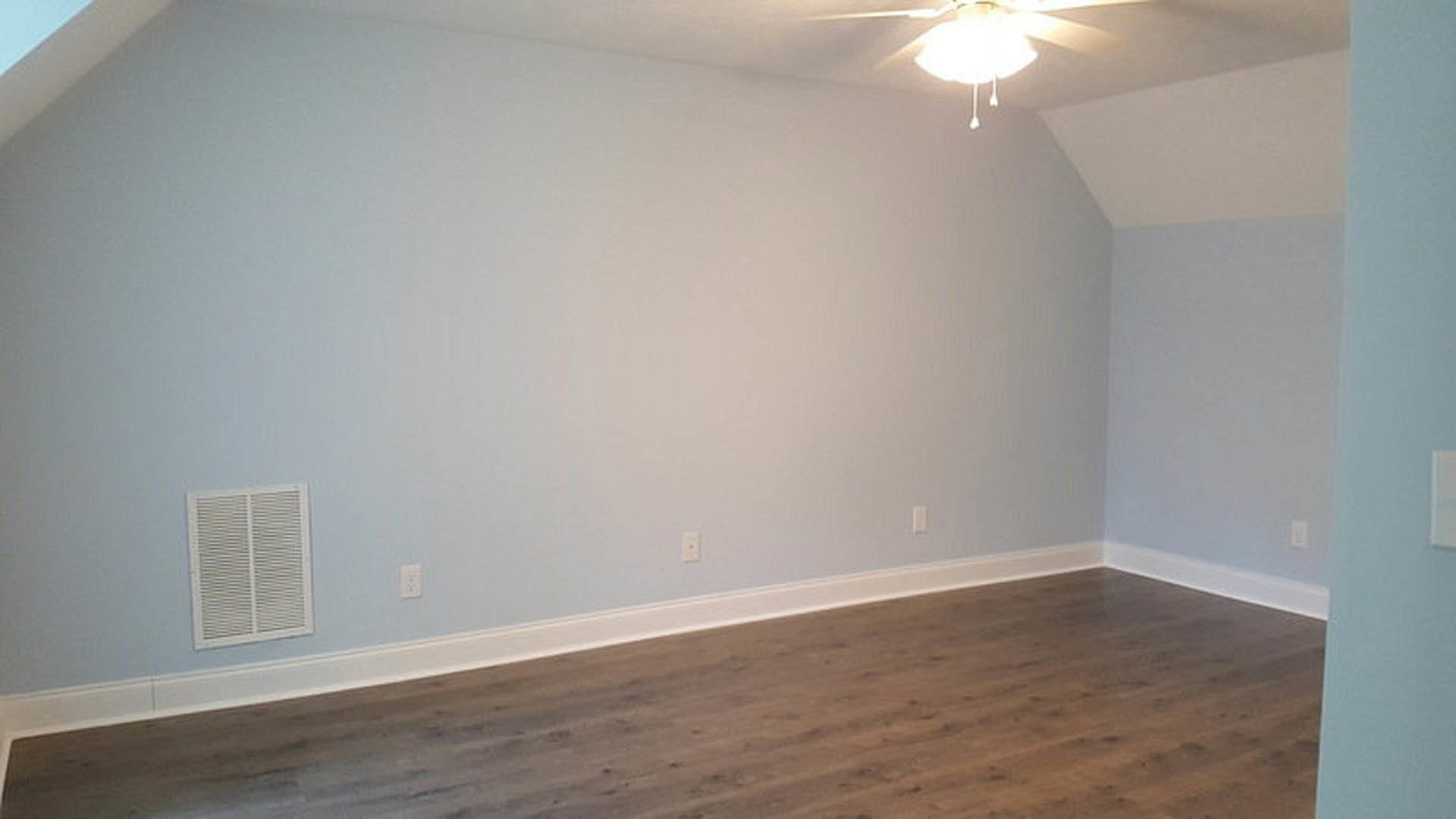 Ceiling fan with light fixture mounted on white plaster ceiling, wood laminate flooring with white baseboards, white wall vent, neutral walls, modern interior design