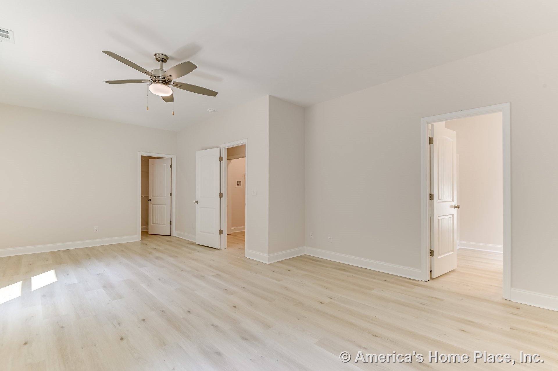 Spacious bedroom with light wood plank flooring, three white paneled doors, ceiling fan with built-in light, neutral walls, white baseboards, and natural light streaming through a