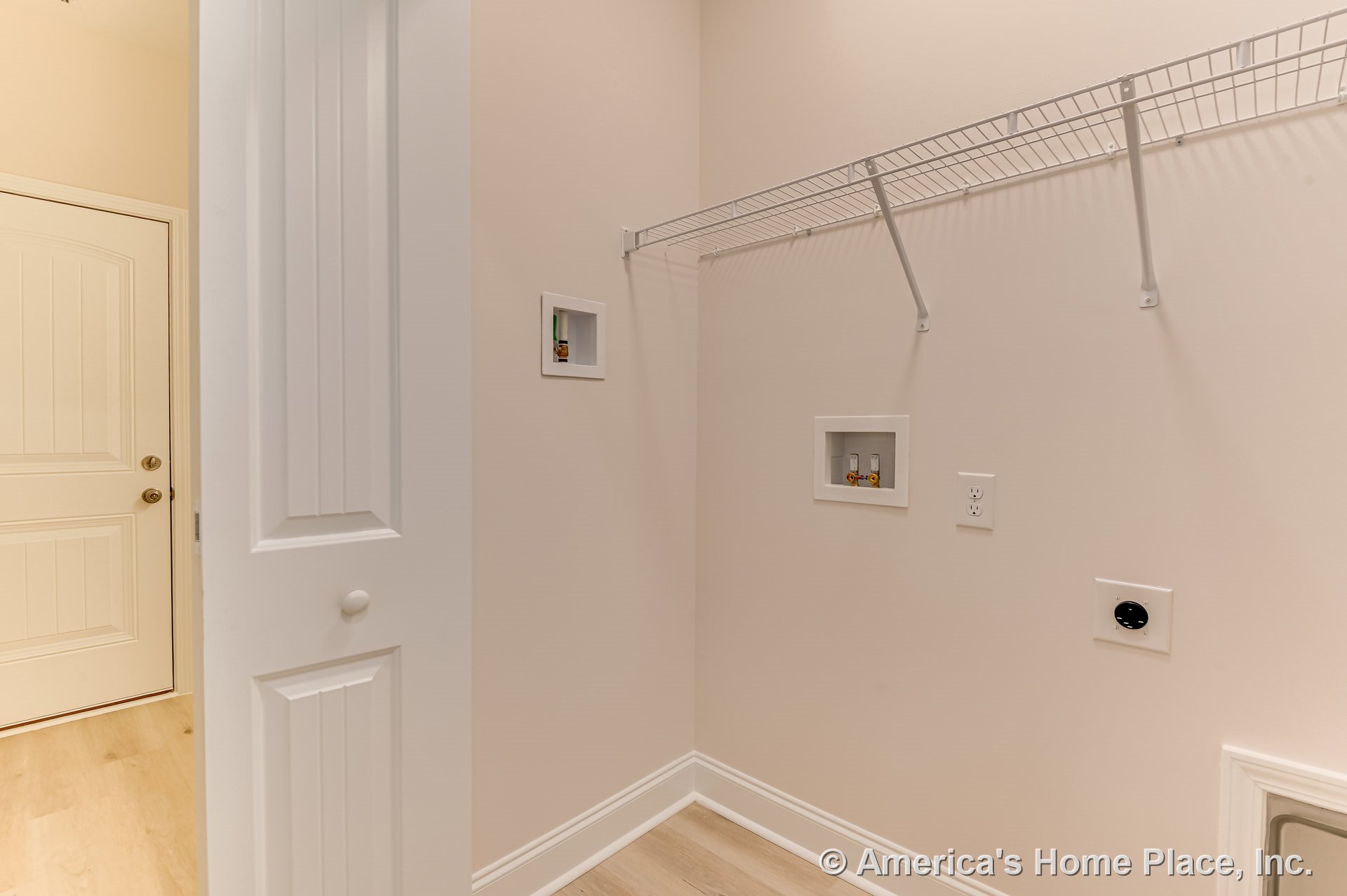 Laundry room with white wire shelf above washer and dryer hookups, recessed utility boxes, light wood plank flooring, white paneled door, baseboard trim, and light-colored walls.