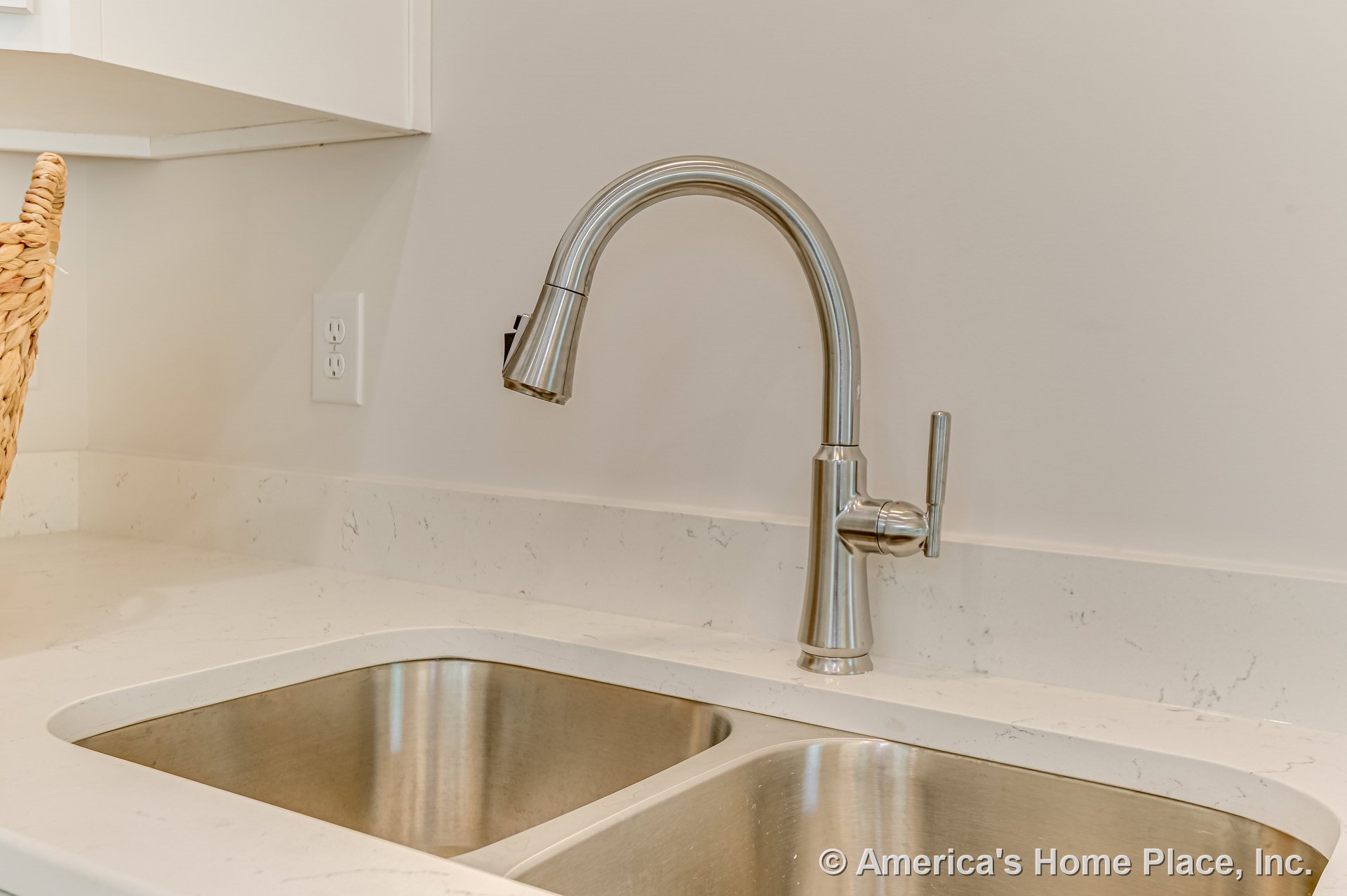 Stainless steel double sink with brushed nickel faucet set in light quartz countertop, white cabinetry, wall outlet above counter, under-cabinet corner.