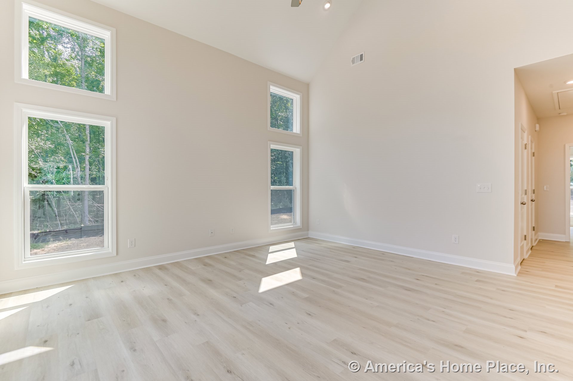 Vaulted ceiling living room with double stacked tall windows, light wood plank flooring, neutral wall paint, white baseboard trim, recessed ceiling lights, and open entry hallway.