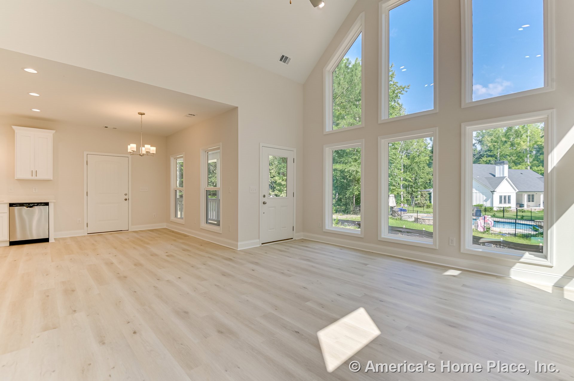 Vaulted ceiling living area with floor-to-ceiling windows, light wood flooring, white trim and baseboards, open floor plan connecting to kitchen with built-in white cabinetry
