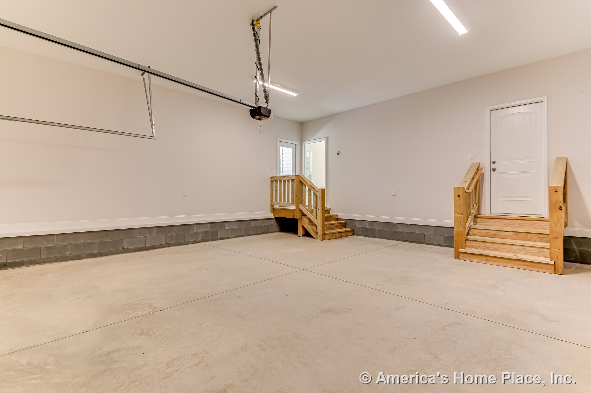 Unfinished garage with exposed concrete floor, two wooden staircases with step railings, white paneled entry door, block foundation wall, garage door opener, recessed ceiling