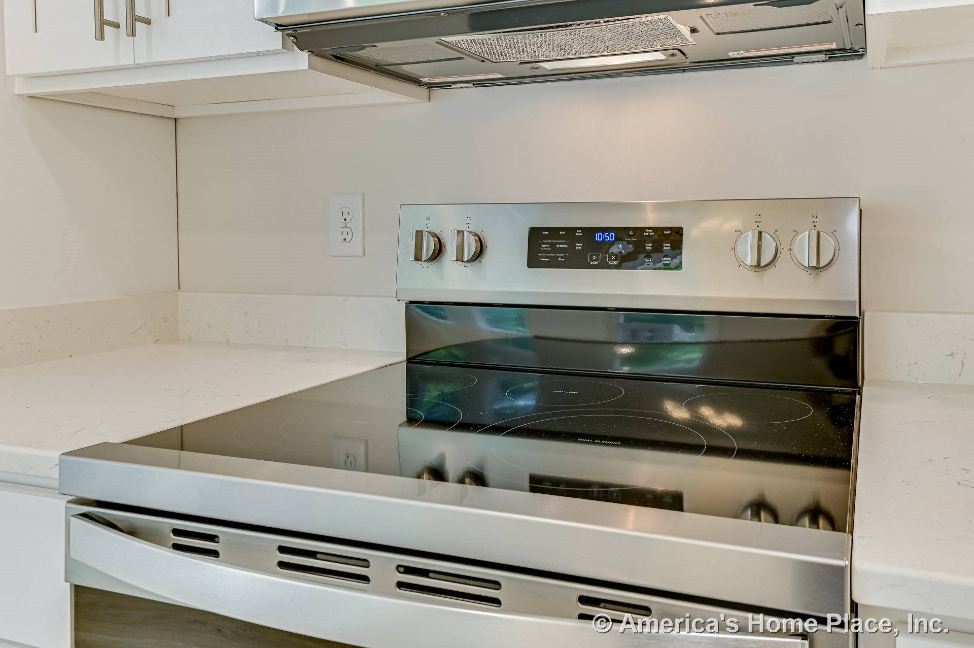 Stainless steel electric range with smooth glass cooktop set against white quartz countertops, white upper cabinetry, built-in range hood, and visible wall outlet.
