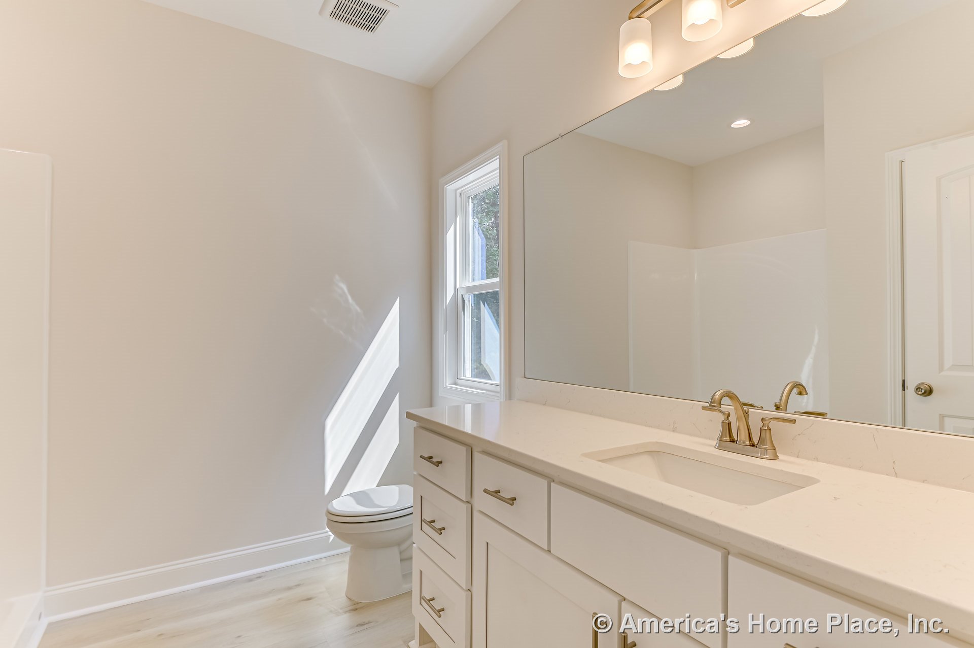 White shaker vanity with quartz countertop and brushed brass faucet, single undermount sink, large wall mirror, three-light brass fixture, window with trim, light wood flooring