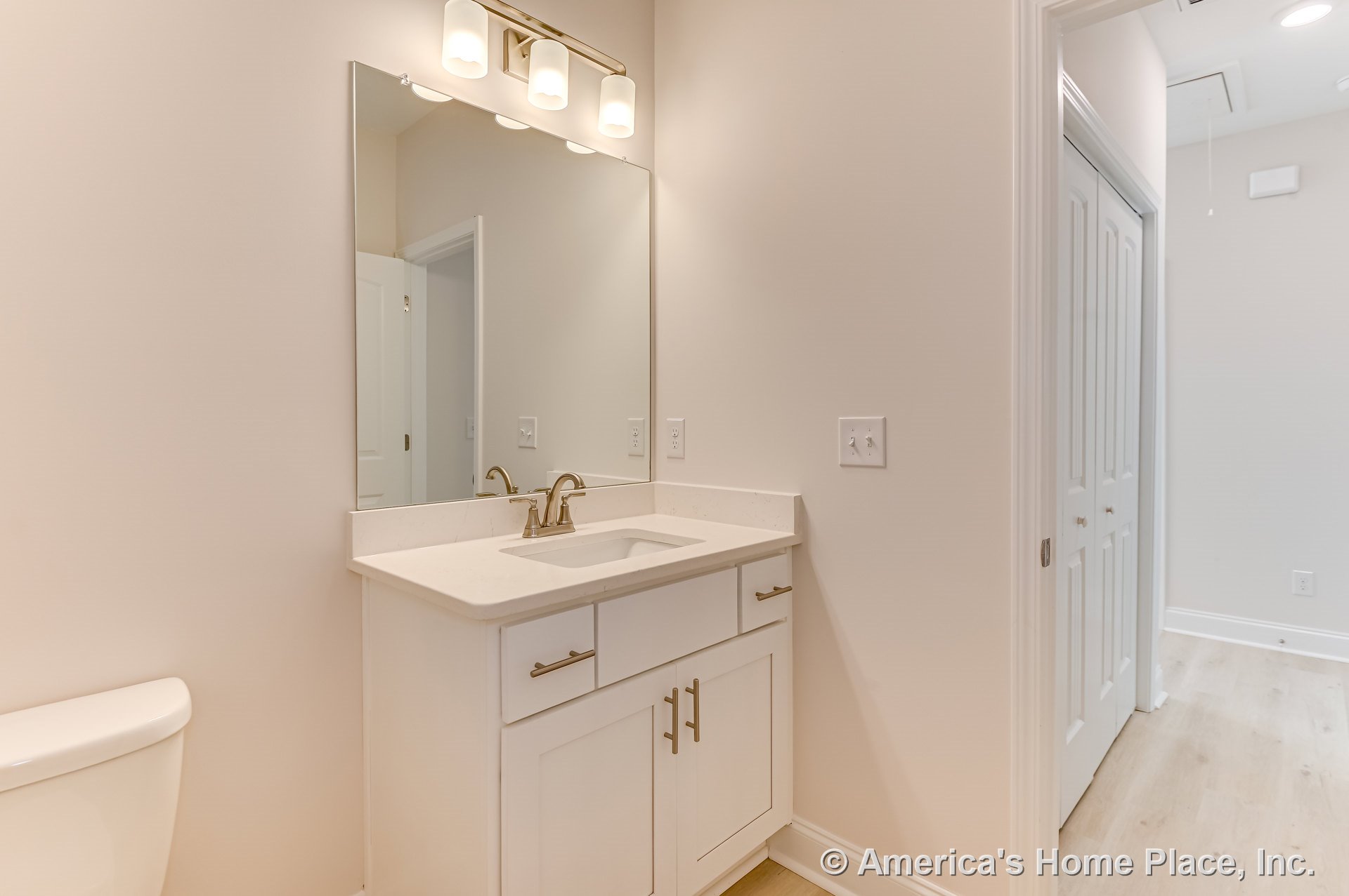 White shaker-style vanity with quartz countertop and brushed nickel faucet beneath a large wall mirror and three-light built-in fixture, light wood flooring, neutral walls, white