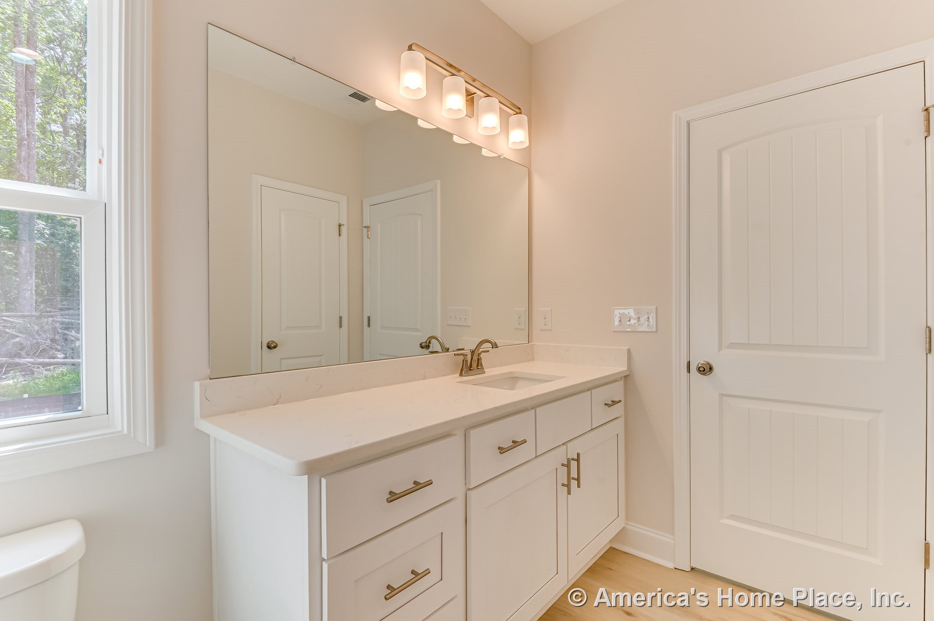 White shaker-style vanity with quartz countertop, brushed nickel faucet, and four-light vanity fixture beneath a large wall mirror; paneled interior door, window trim, neutral