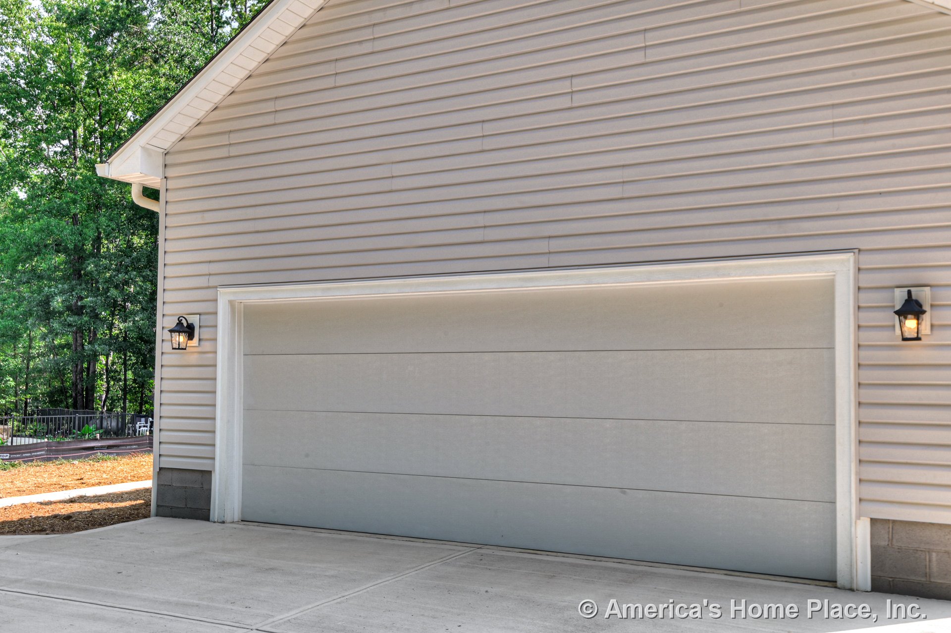 Double garage door with white trim set in vinyl siding exterior, concrete driveway, gable roof edge, and two wall-mounted lantern lights flanking the entrance.