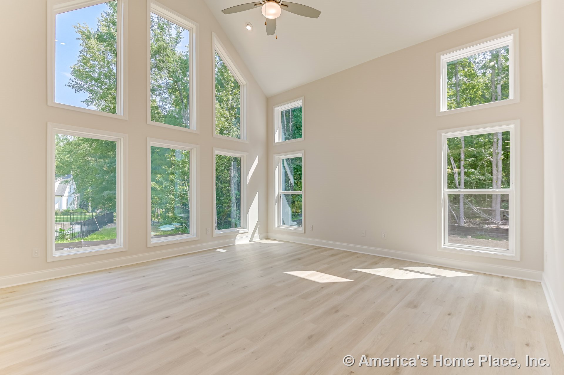 Vaulted ceiling living room with multiple tall picture windows, light wood flooring, ceiling fan with light fixture, white window trim, and neutral wall color.