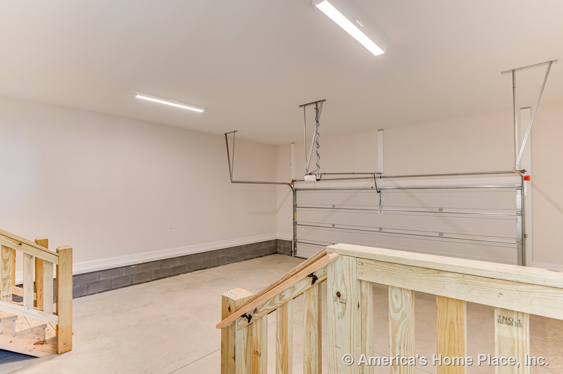 Double overhead garage door with concrete flooring, unfinished wood stairs and railing, block foundation wall, fluorescent ceiling lights, and neutral painted walls.