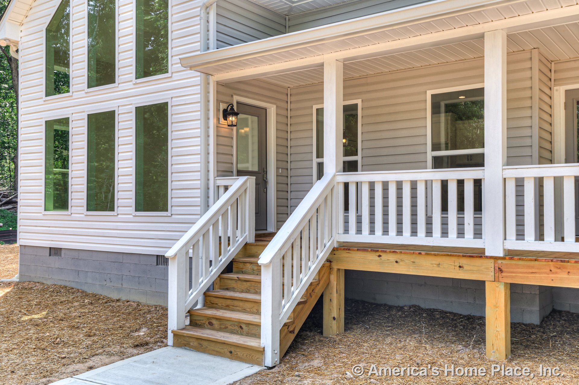 Covered front porch with white railings and wooden steps, multiple tall vertical windows, vinyl siding, exterior light fixture near front door, concrete walkway, and overhang above