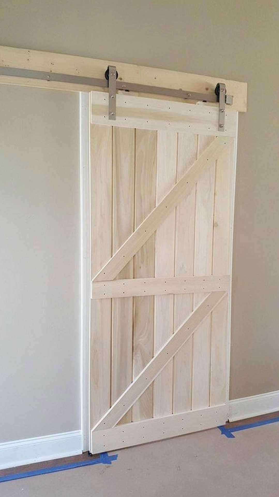 White hinged door next to rustic wood sliding barn door with cross-brace detail, wood handle, blue painter’s tape on adjacent wall, exposed wood beam overhead, white wall with