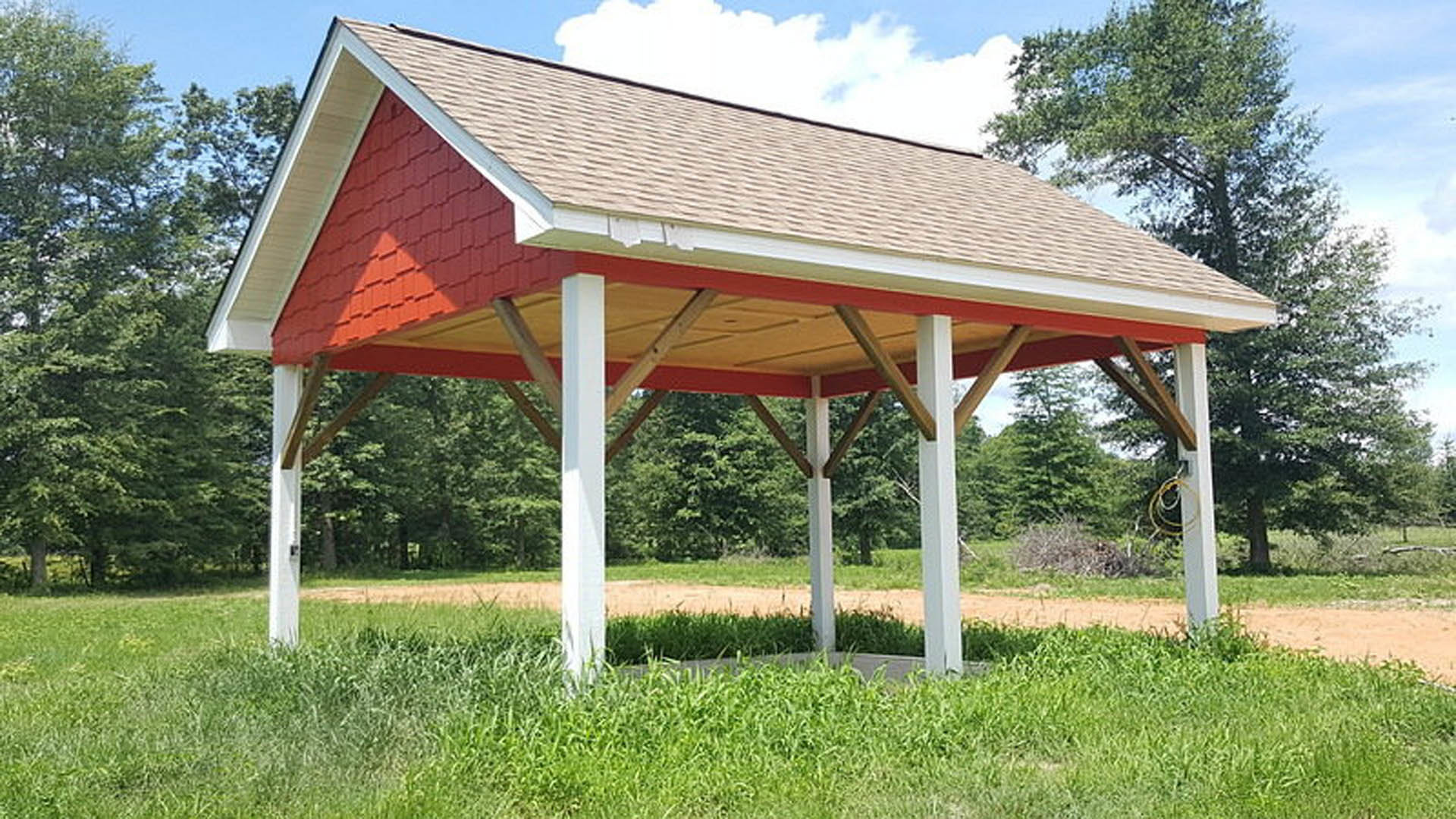 Red and white pavilion with open sides set on a grassy lawn, surrounded by trees and plants under a blue sky