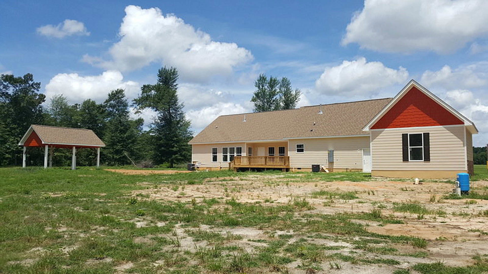 Red roof farmhouse with white pillars and wooden porch railing, surrounded by a large grassy yard, white-framed windows, and mature trees under a partly cloudy sky.