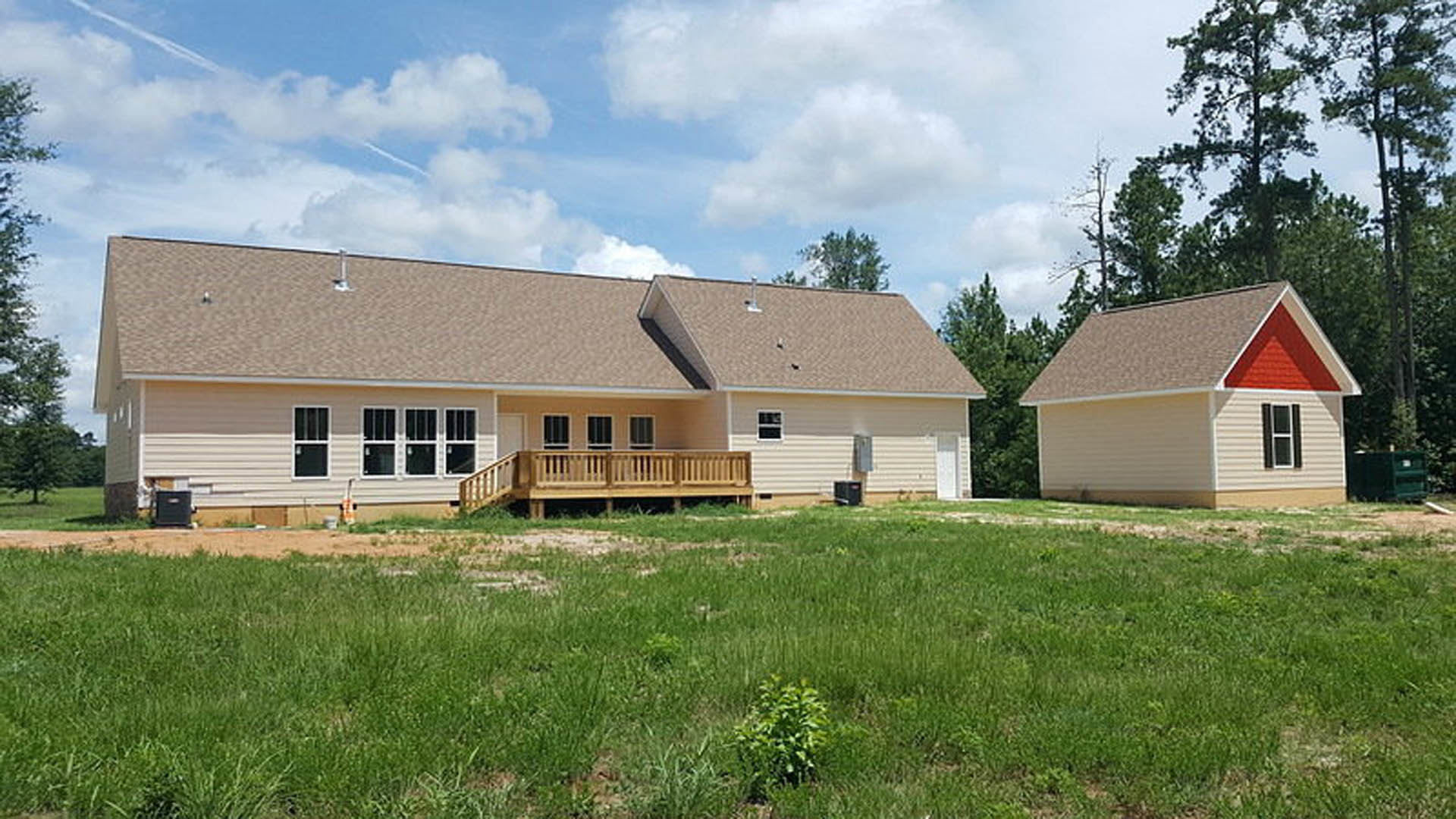 Two-story house with red roof, white-framed windows, wooden porch and deck with railing, surrounded by green lawn and small plants