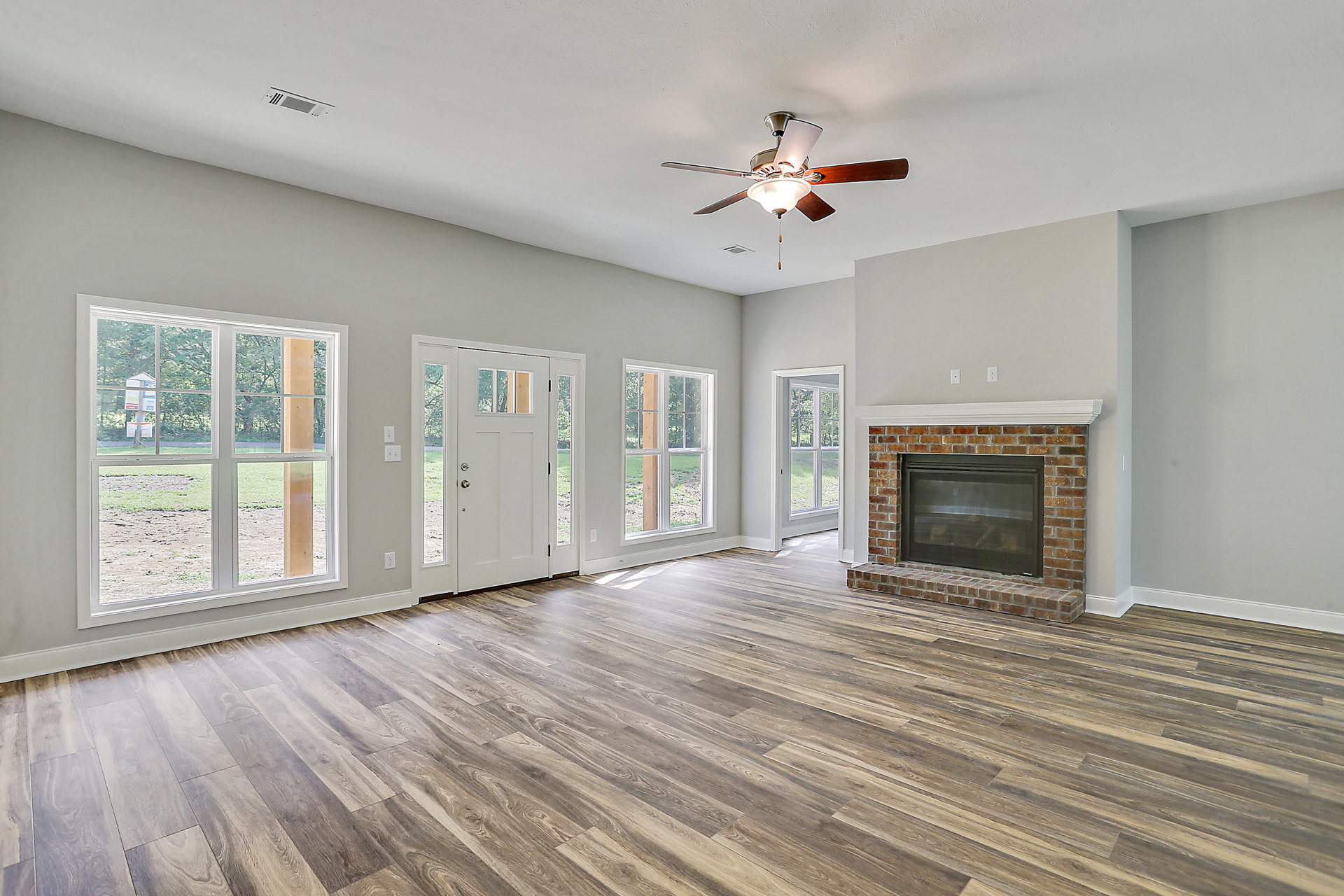 Living room with wood flooring, brick hearth fireplace with glass door, ceiling fan with light fixture, white doors, and window showing sign outside