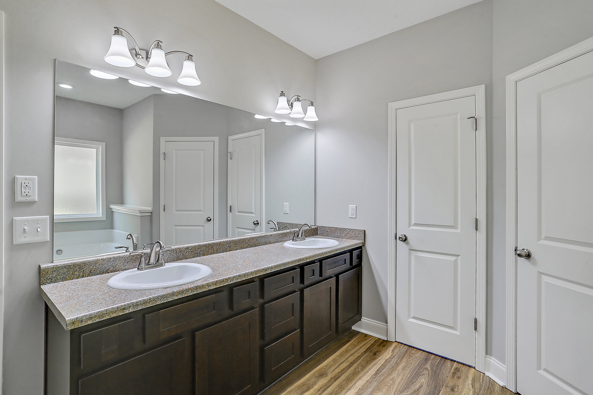 Bathroom with two undermount sinks set in a quartz countertop, wide frameless mirror above, white shaker cabinets, chrome faucets, and modern light fixture with white shade.
