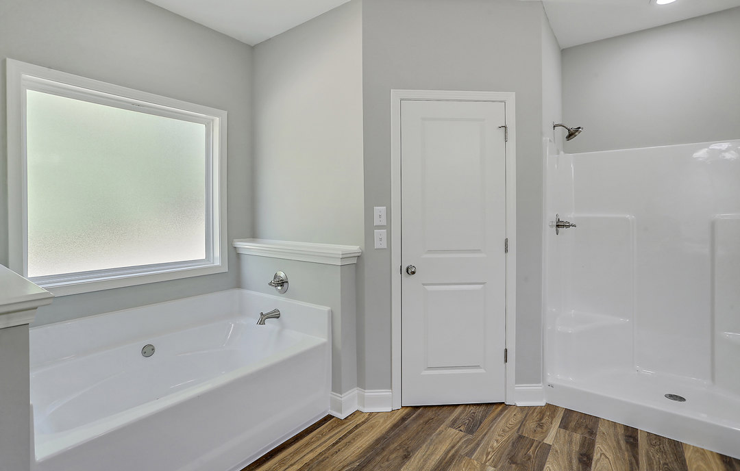 White bathtub with silver faucets beside frosted glass window, white door with silver knob, wood flooring, and white shower with silver faucet