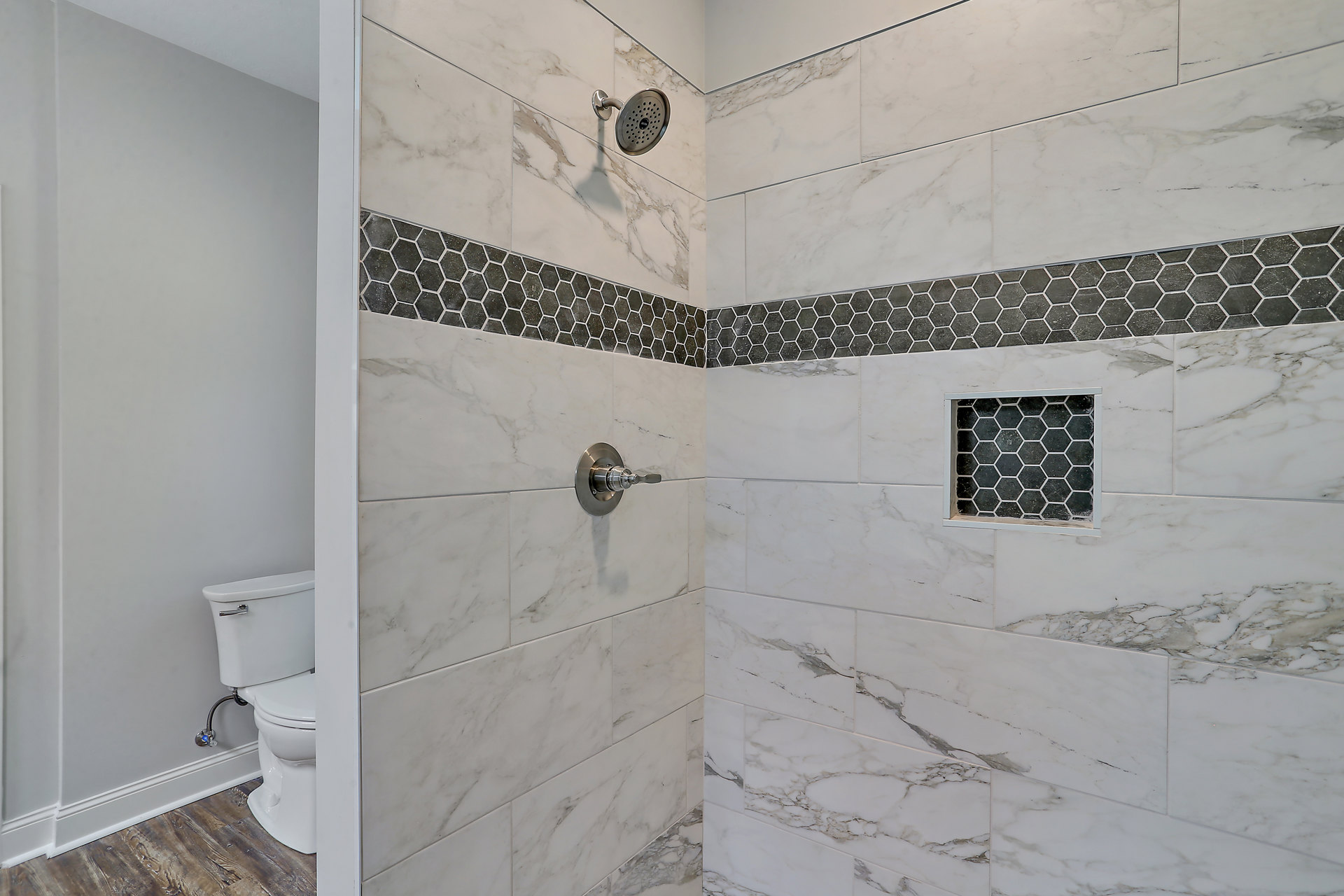 Marble tile shower with circular metal shower head, white toilet with metal hose, white square window with hexagon pattern, close-up of chrome faucet