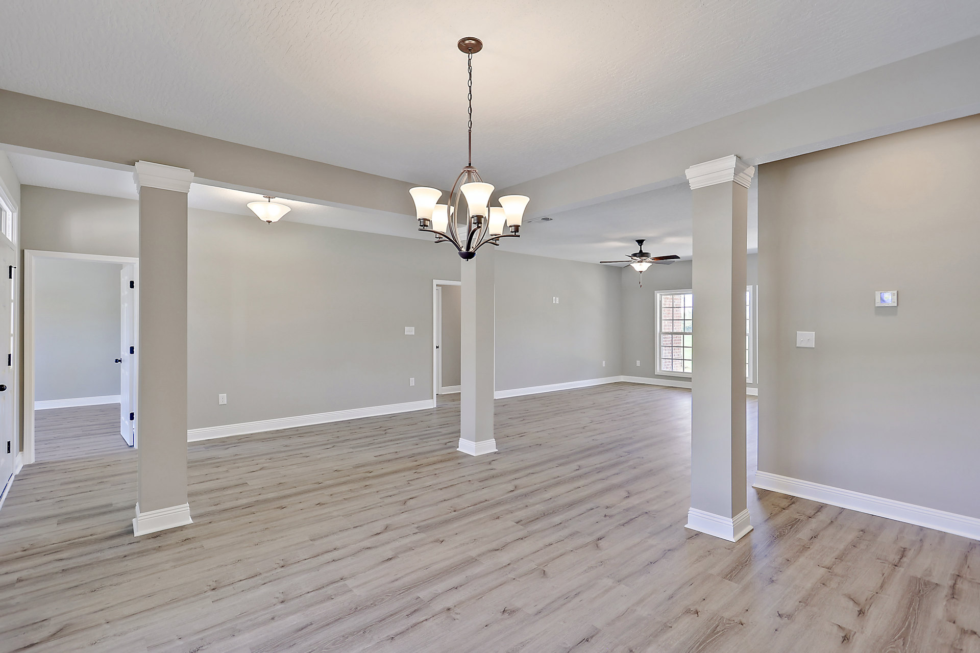 Open living space with wood plank flooring, ceiling fan with light fixture, white columns, neutral plaster walls, and large window framed by exposed brick.