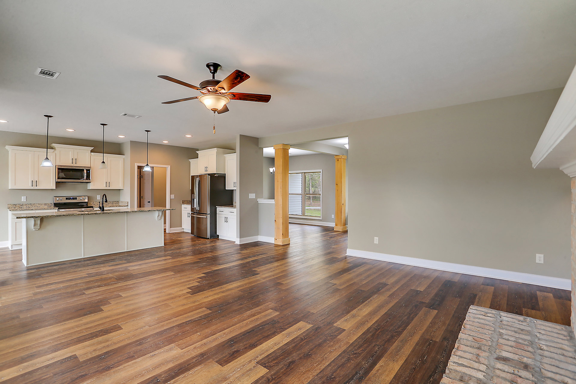 Open living room with wood flooring, white walls, ceiling fan with light fixture, adjacent kitchen featuring stainless steel refrigerator, built-in microwave, and white cabinetry.