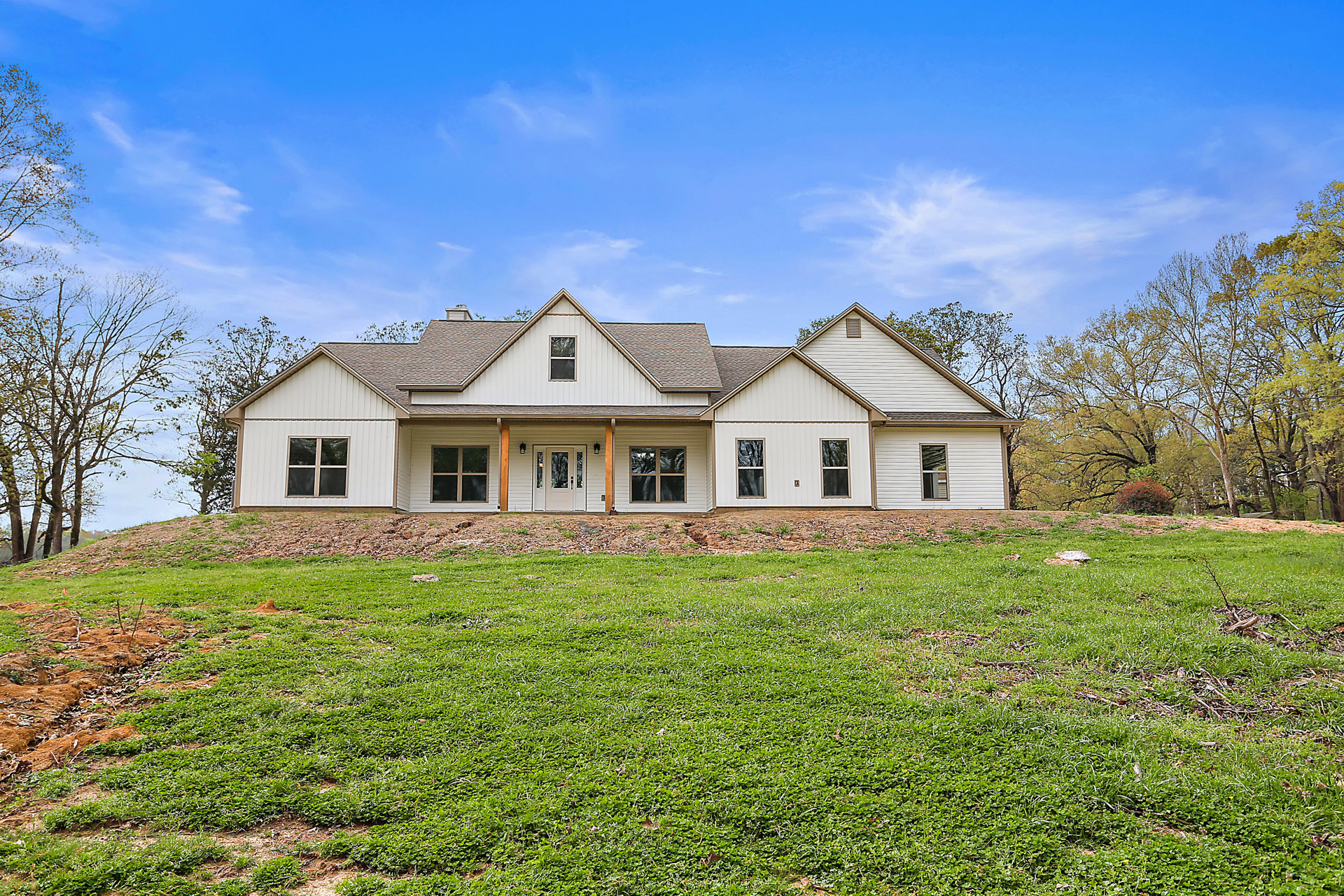 Two-story farmhouse with white siding, brown-framed windows, and a white front door, surrounded by green lawn and mature trees, blue sky overhead, Robert Frost Farm visible in the