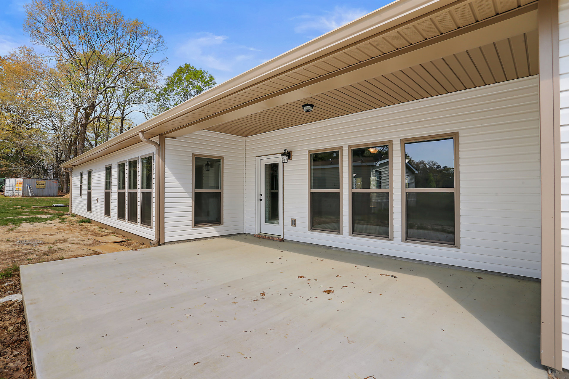 Two-story home with gray siding, covered front porch, white door with glass window, concrete patio scattered with leaves, large windows reflecting trees and cloudy sky