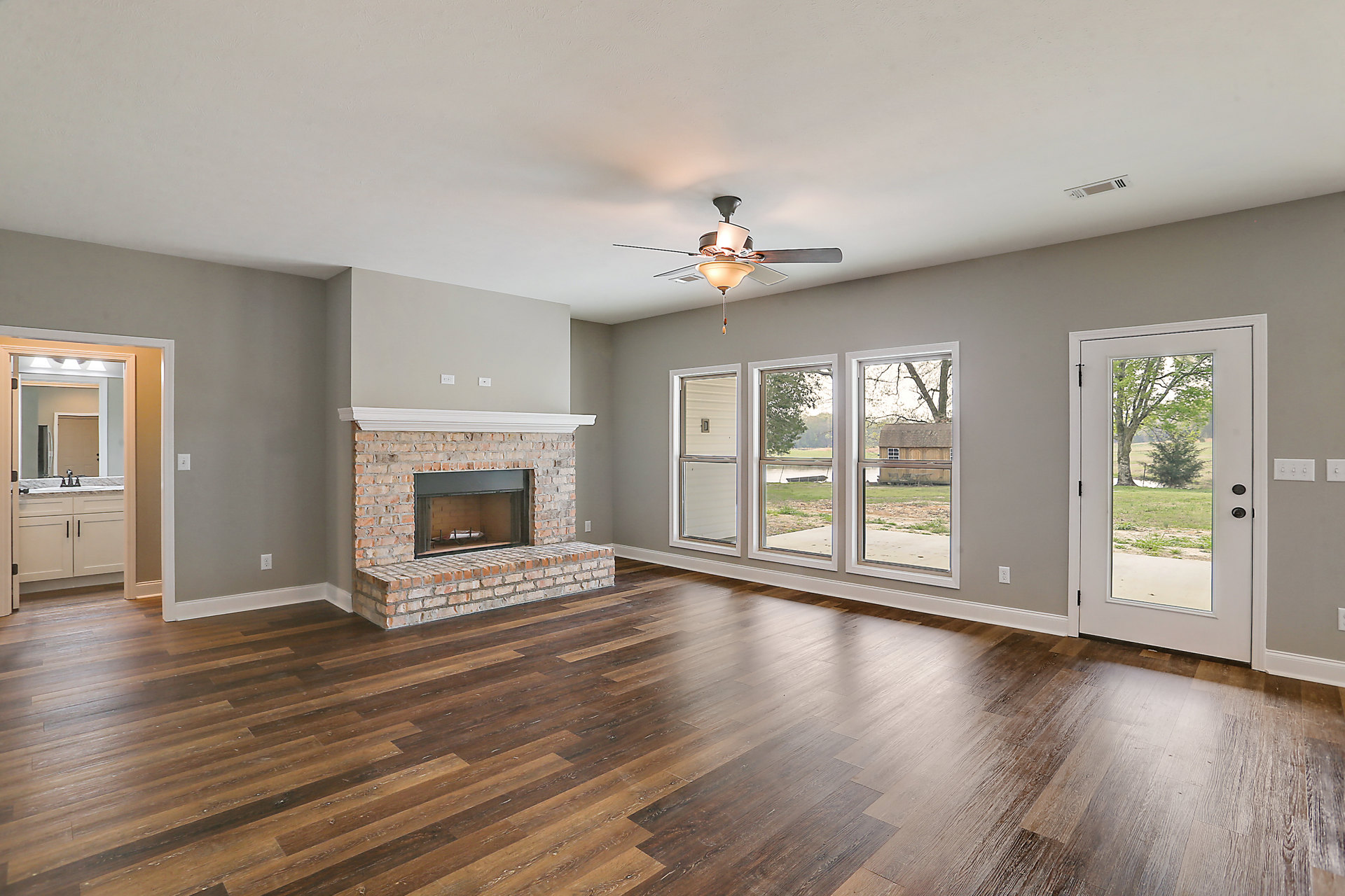 Living room with hardwood floors, brick fireplace featuring a grill, ceiling fan with light fixture, glass door revealing a tree outside, and windows along the wall.