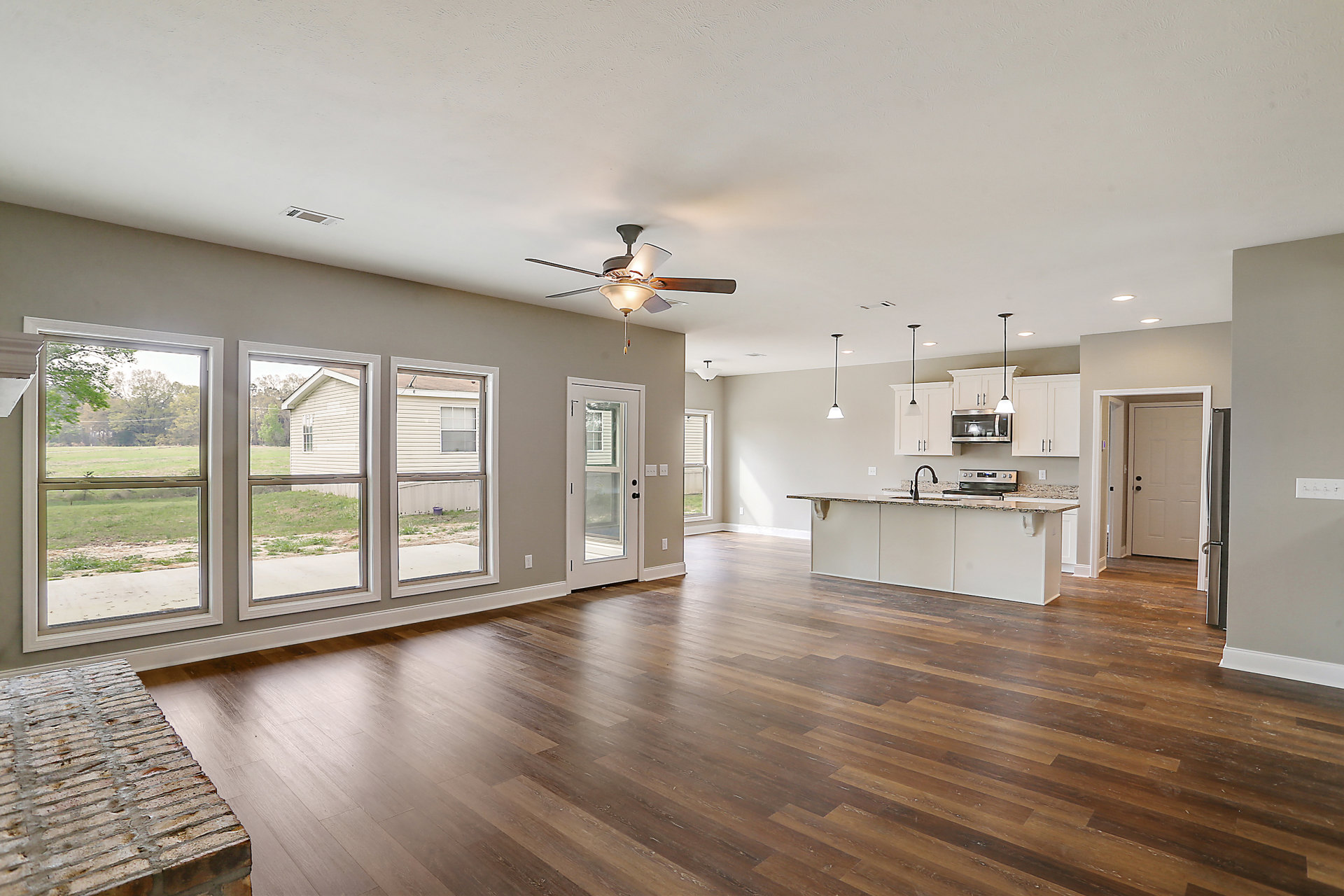 Spacious open floor plan featuring a kitchen and dining area, wood flooring, brick dining table, ceiling fan with light fixture, glass doors and windows overlooking grass and